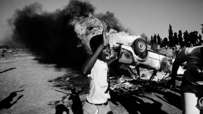 As a car bus behind him, a young South African participates in a civil disturbance outside the Auduza Cemetery.