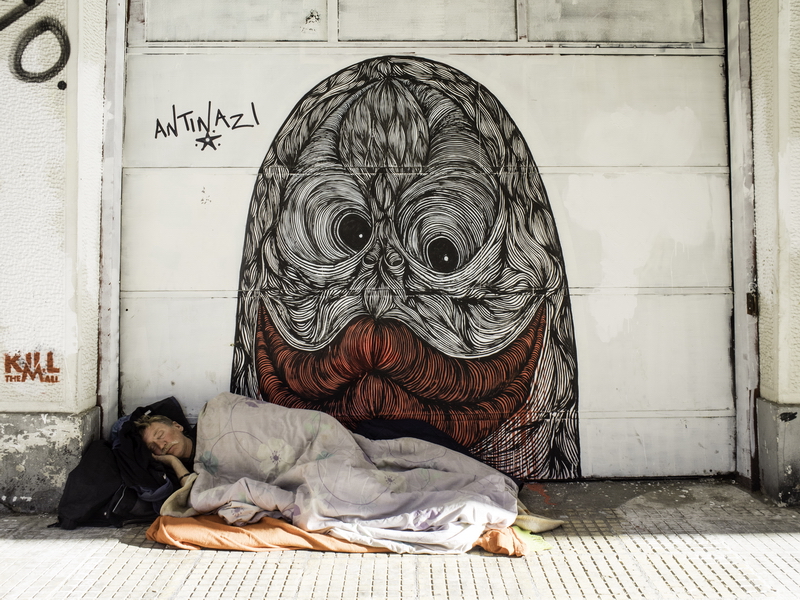 Ein Obdachloser Mann schläft vor einer Graffitimalerei, die ein Monster mit grossem roten Mund zeigt. Das Monster ist aus vielen Linien aufgebaut.