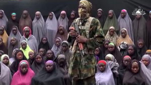 This video grab image created on August 14, 2016 taken from a video released on youtube purportedly by Islamist group Boko Haram showing what is claimed to be one of the groups fighters at an undisclosed location standing in front of girls allegedly kidnapped from Chibok in April 2014. Boko Haram on August 14, 2016 released a video of the girls allegedly kidnapped from Chibok in April 2014, showing some who are still alive and claiming others died in air strikes. The video is the latest release from embattled Boko Haram leader Abubakar Shekau, who earlier this month denied claims that he had been replaced as the leader of the jihadist group. / AFP PHOTO / HO / RESTRICTED TO EDITORIAL USE - MANDATORY CREDIT "AFP PHOTO / BOKO HARAM" - NO MARKETING NO ADVERTISING CAMPAIGNS - DISTRIBUTED AS A SERVICE TO CLIENTS